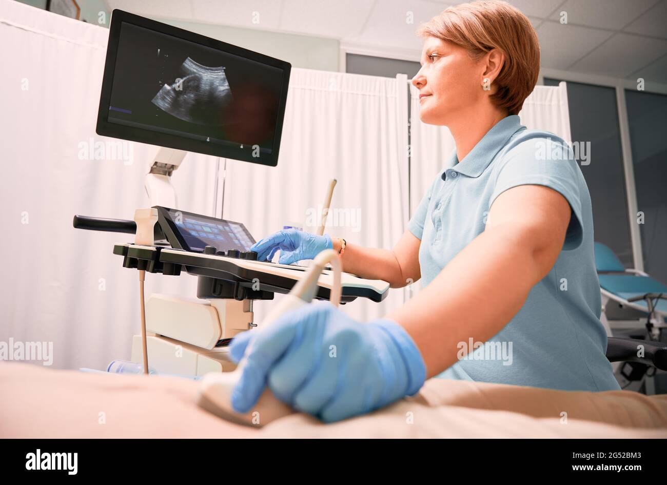 Donna ecografa che esamina donna con ecografo. Il medico sposta il trasduttore a ultrasuoni sull'addome del paziente mentre guarda il display. Concetto di esame medico e diagnostica ecografica. Foto Stock