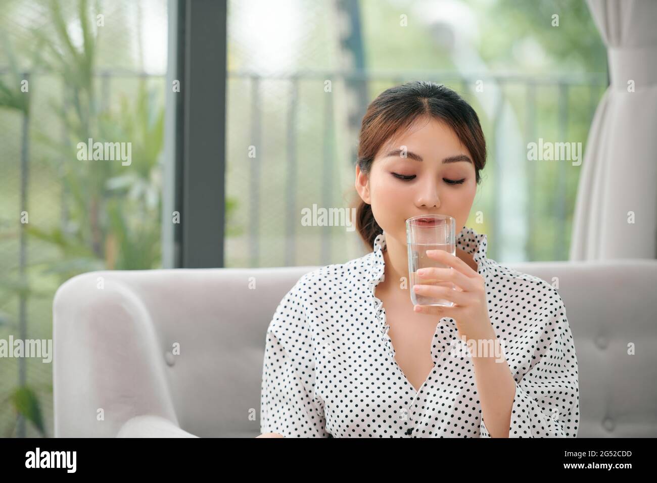 Donna acqua potabile a casa e guardare è seduta su un divano nel salotto di casa Foto Stock