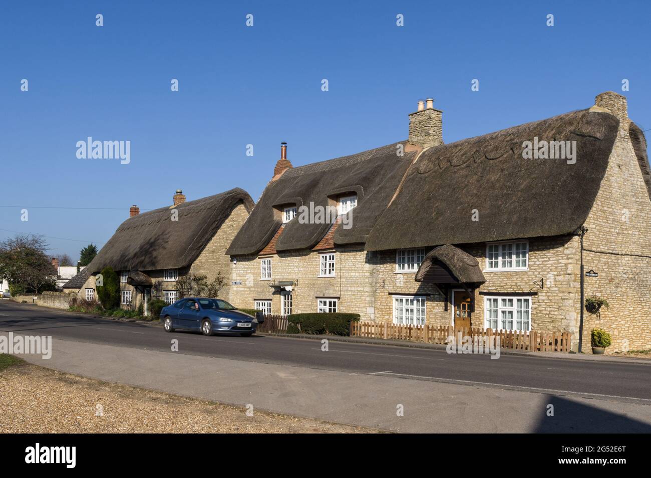 Scena di strada in primavera nel villaggio di Stoke Goldington, Buckinghamshire, Regno Unito; auto che passa una fila di case di paglia. Foto Stock