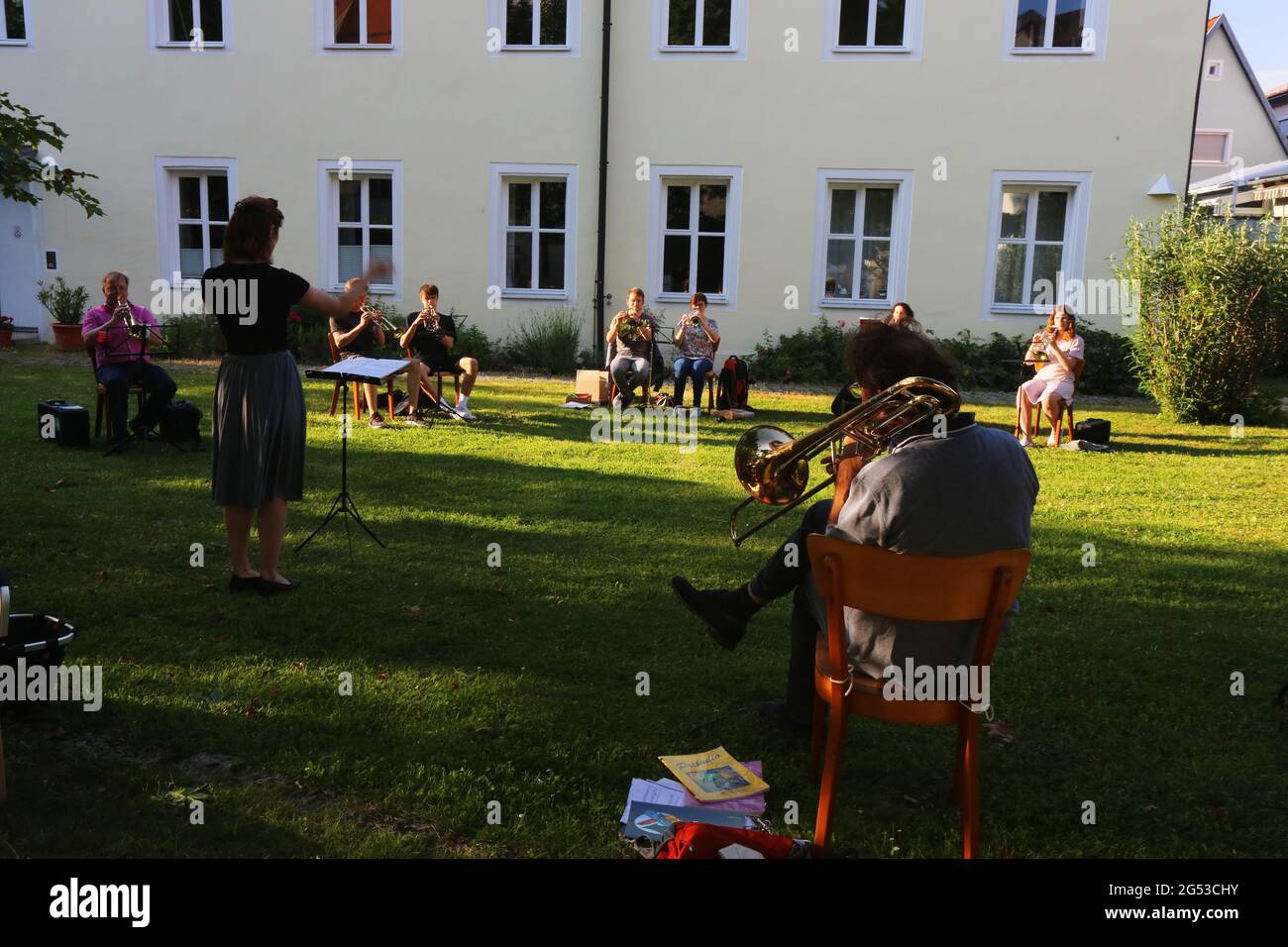 Ein Bläserensemble probt im schattigen Garten eines alten Gebäudes neue Musikstücke mit Trompete, Posaune und Tuba unter Anleitung einer Dirigentin. Foto Stock