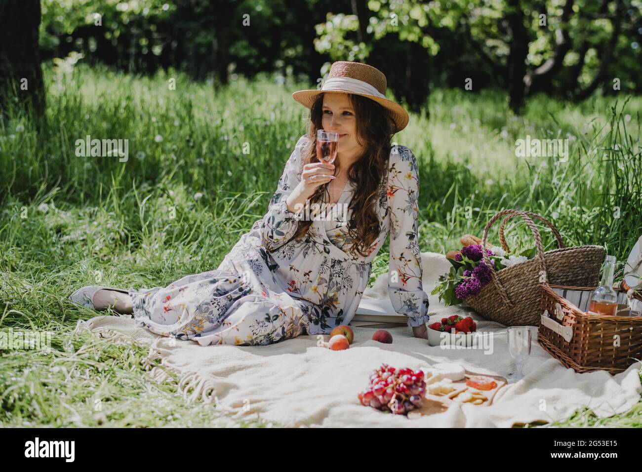Giovane donna in abito estivo e cappello di paglia seduta con vino rosato, formaggio, frutta fresca e baguette su coperta. Foto Stock