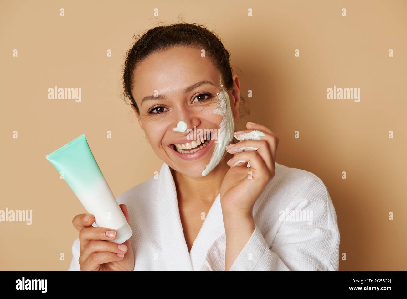 Primo piano ritratto di bellezza di una giovane donna con bel sorriso toothy applicando una crema facciale o scrub sul viso in piedi isolato su sfondo beige Foto Stock