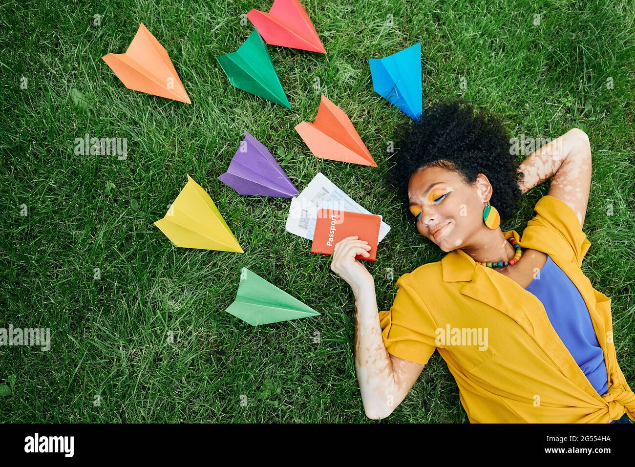 Concetto di viaggio. Donna multietnica con biglietti aerei e passaporto internazionale in mano sogni di vacanze sdraiati su erba verde intorno a pape colorata Foto Stock