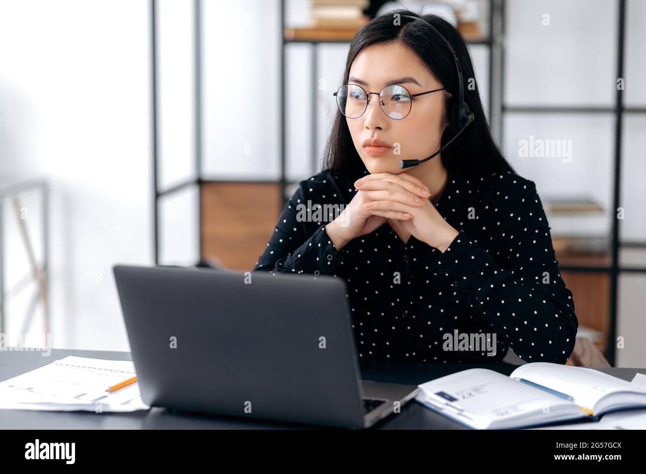 Giovane donna bruna asiatica sconvolto con occhiali e una cuffia, un operatore di supporto o manager, seduto a una scrivania, guardando da parte pensieroso, infelice, con stress, ha bisogno di riposo, preoccupato Foto Stock