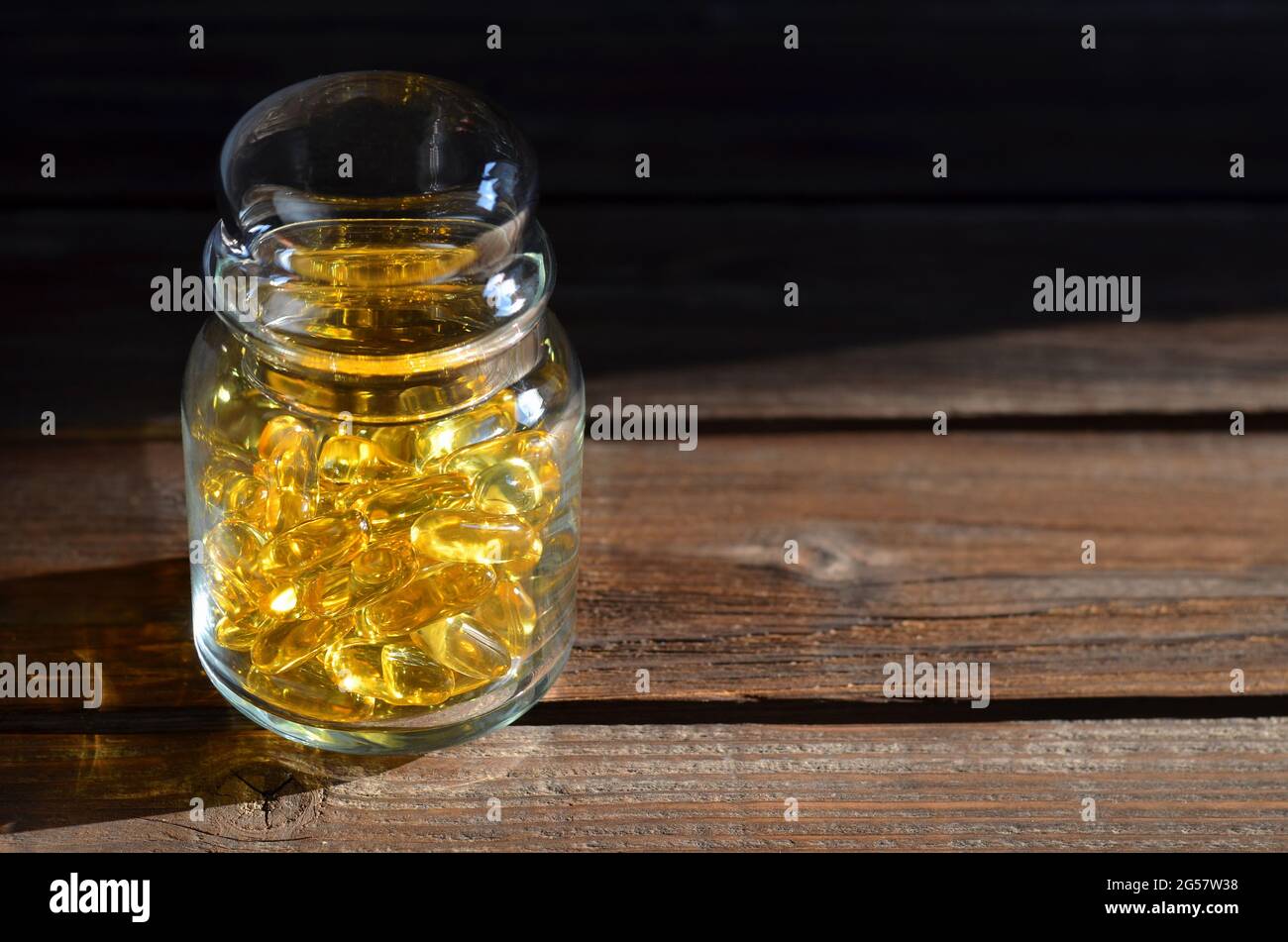 Vaso di vetro con integratori alimentari in capsule gialle trasparenti illuminate dalla luce solare su un vecchio tavolo di legno. Vitamine per immunità e longevità. Foto Stock
