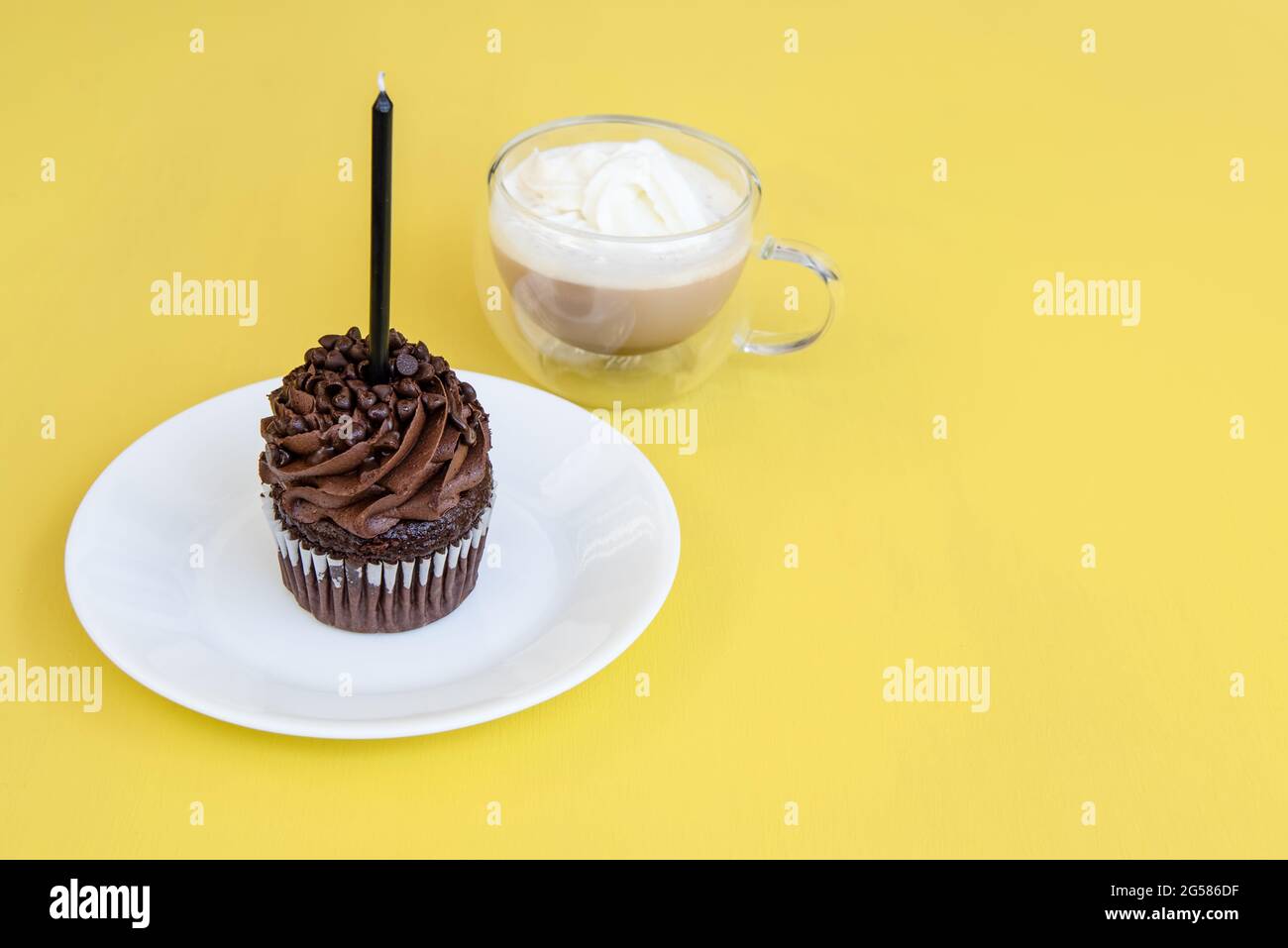 Disposizione piatta di un cupcake al cioccolato con una candela nera e una tazza di cappuccino su sfondo giallo. Foto Stock