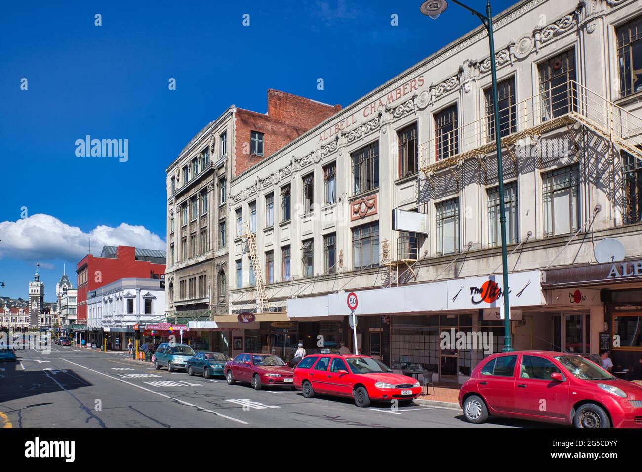 Scena di strada con negozi frontali e automobili che guardano verso la stazione ferroviaria in lontananza, a Dunedin, Isola del Sud, Nuova Zelanda Foto Stock