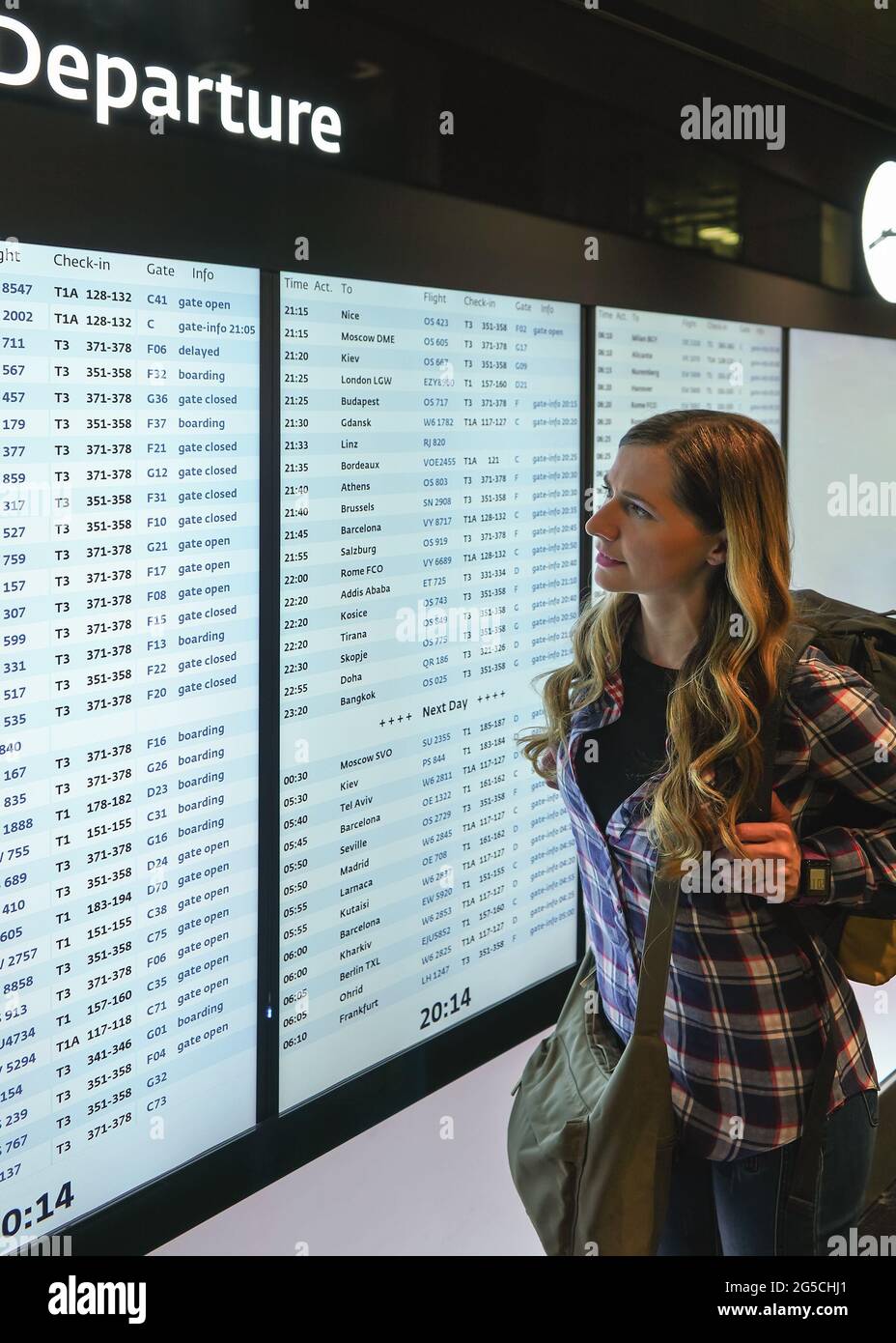 Giovane donna con zaino verde e camicia che guarda lo schermo del bordo di partenza all'aeroporto Foto Stock