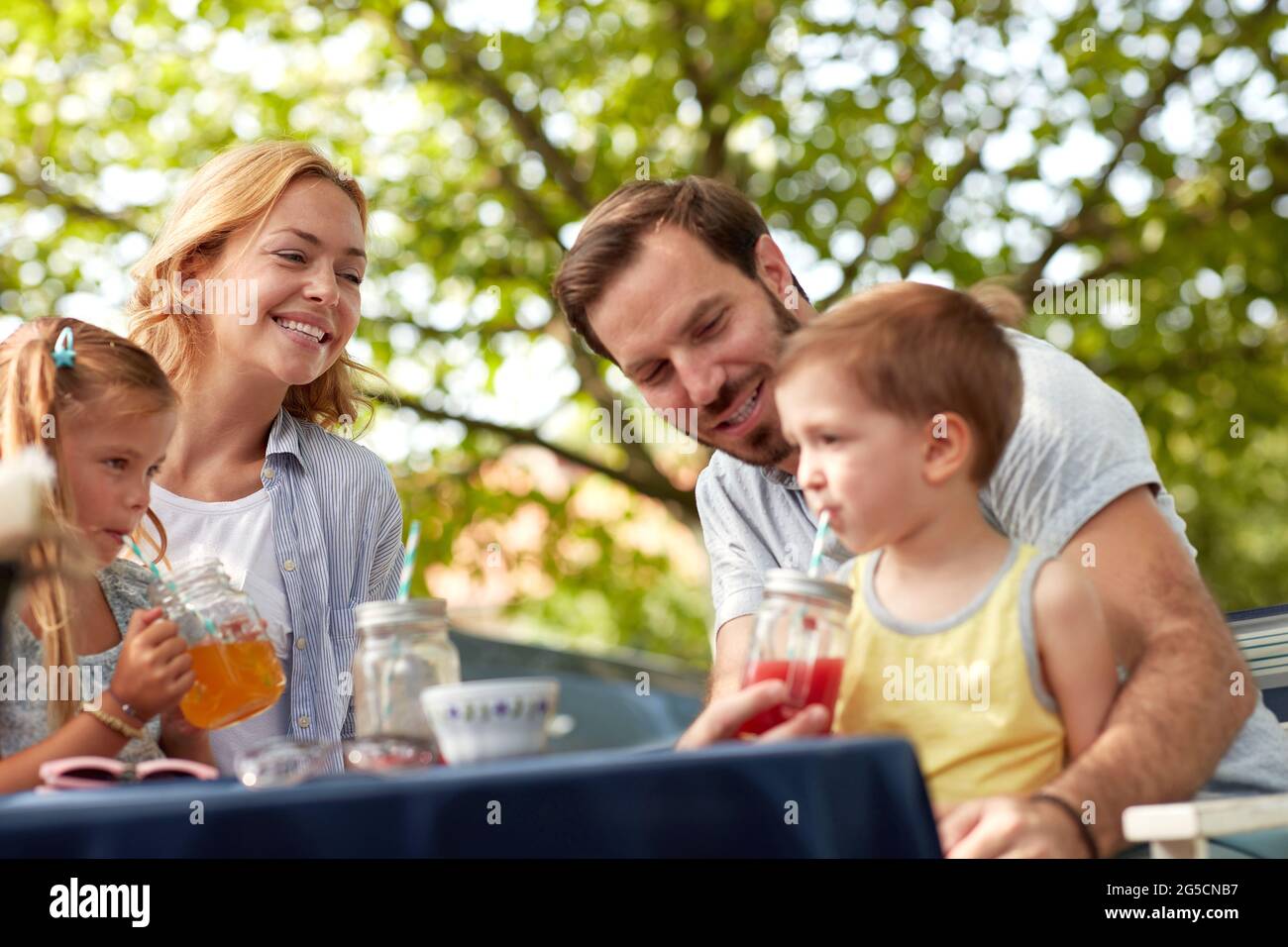 allegra famiglia caucasica seduta all'aperto, bevendo succhi, godendosi. padre aiutare suo figlio a bere succo sulla paglia. Foto Stock