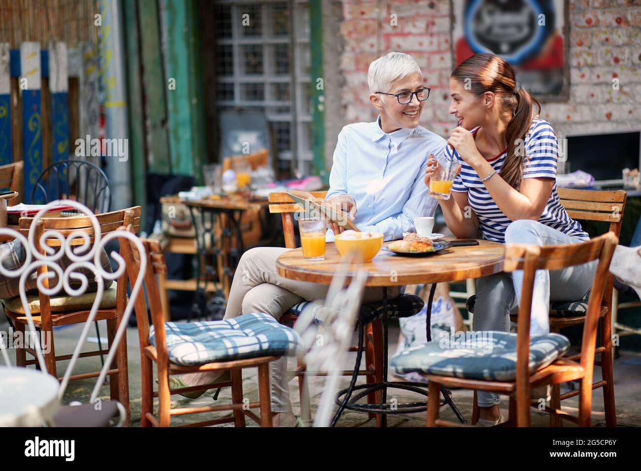 donna anziana che mostra parte nel libro aperto alla giovane donna bruna adulto che beve succo d'arancia su una paglia, seduto in un caffè all'aperto. Insegnante e favori Foto Stock