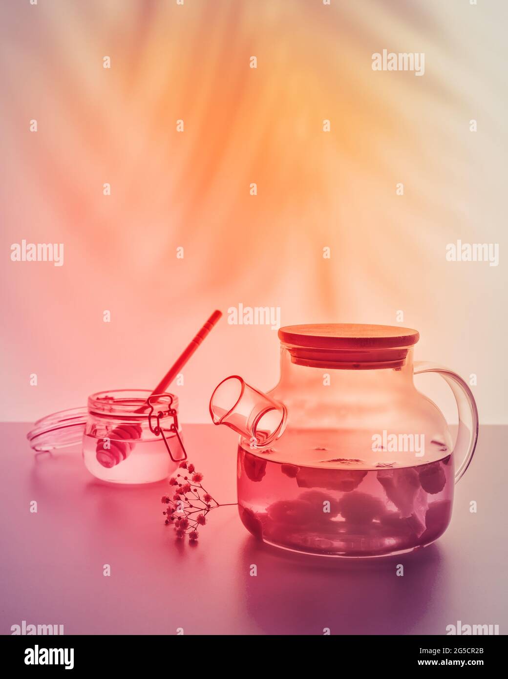 Estate, relax tè tempo. Teiera in vetro con tè alla frutta e vasetto con miele su uno sfondo chiaro e tramonto con ombre lasciate dalle palme. Estate, bevande salutari Foto Stock