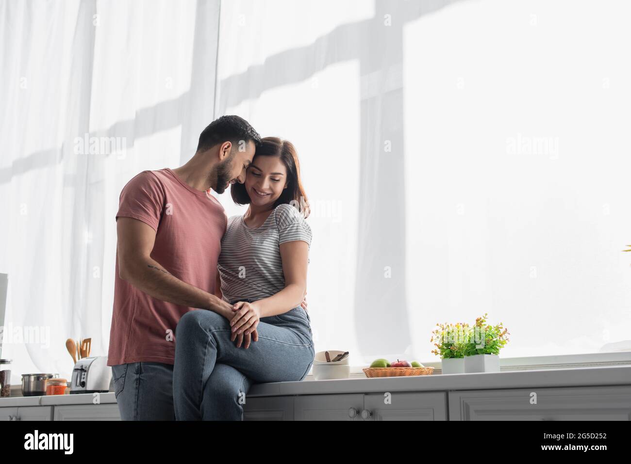 giovane uomo sorridente abbracciando la fidanzata con la mano sull'anca in cucina Foto Stock