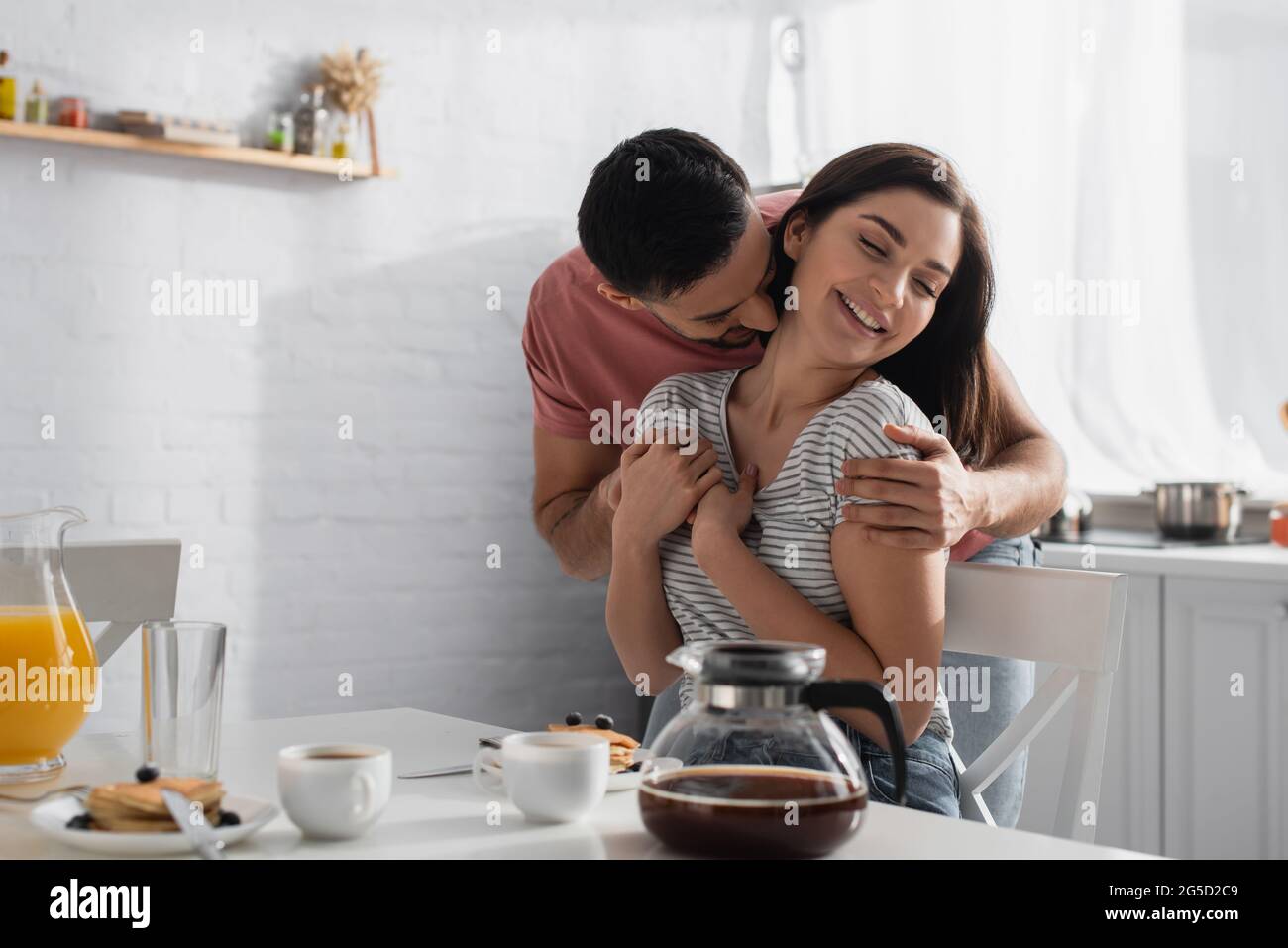 felice giovane coppia abbracciando e baciando vicino al tavolo con colazione, caffè e succo d'arancia in cucina Foto Stock