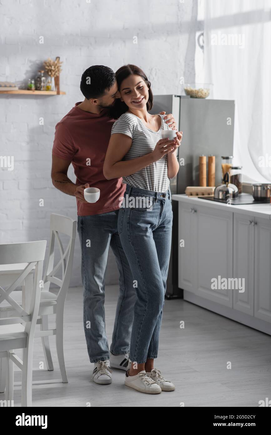 sorridente giovane coppia abbracciando e baciando con tazze da caffè vicino a tavolo e sedie in cucina Foto Stock