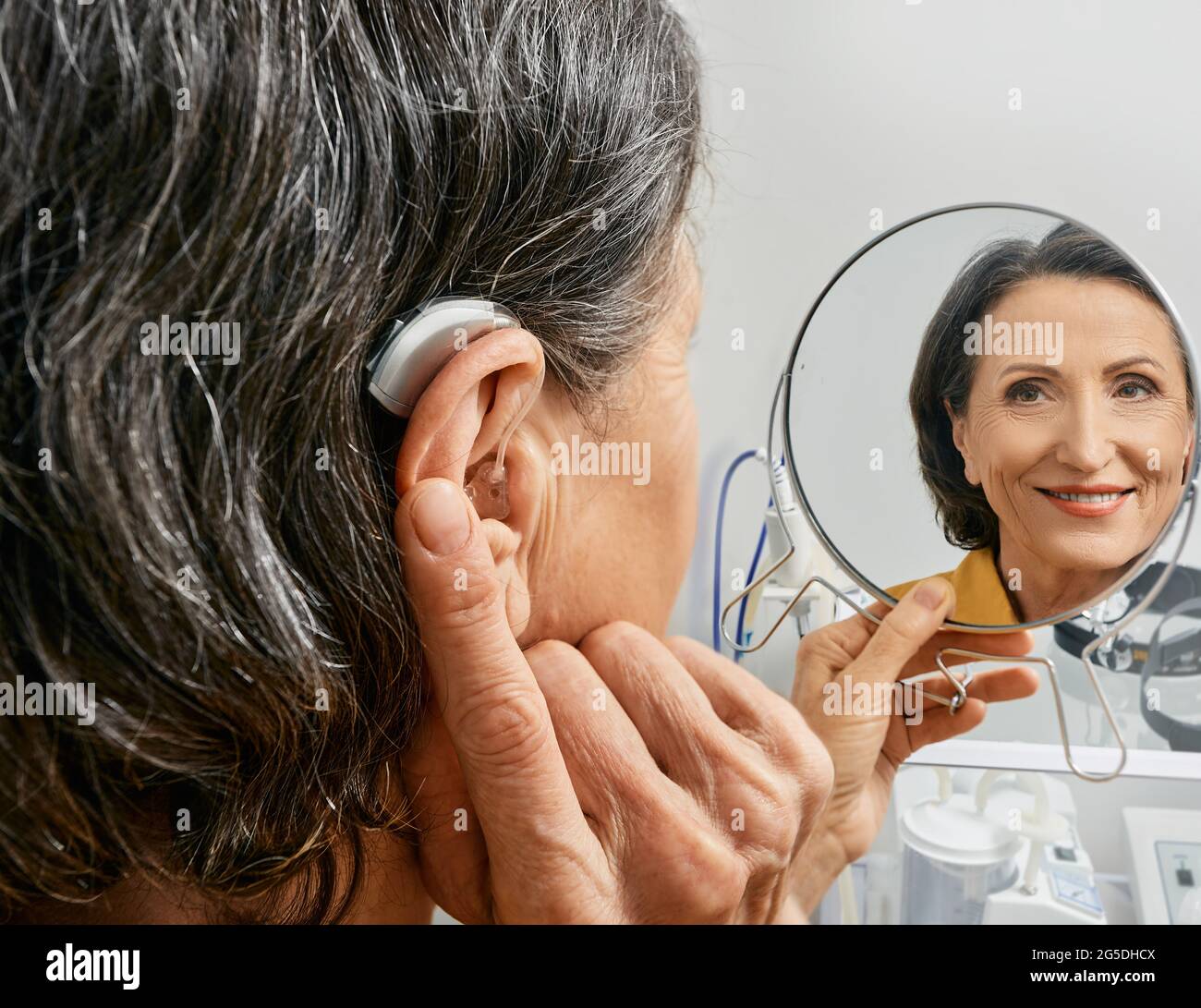 Donna matura con apparecchio acustico BTE guarda se stessa nello specchio e cerca di udire dispositivo, primo piano. Trattamento per perdita dell'udito Foto Stock