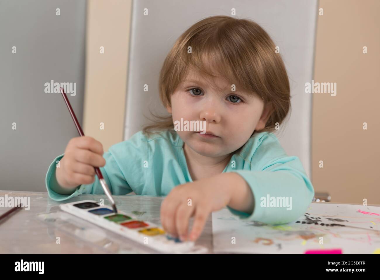 Una ragazza di tre anni in una blusa turchese al tavolo disegna con vernici acquerello. Guarda la fotocamera. Verticale Foto Stock