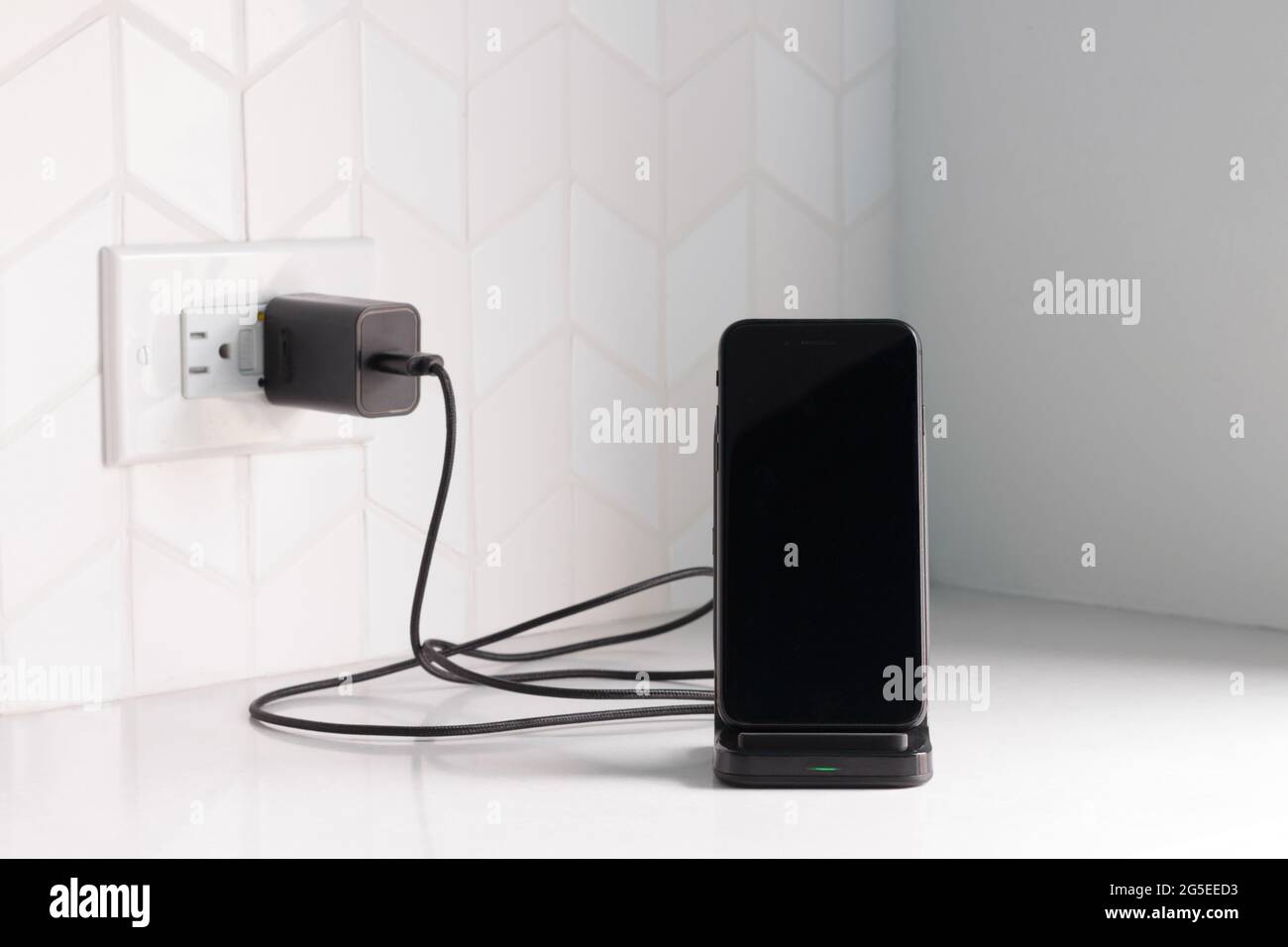 uno smartphone nero inserito in una stazione di ricarica wireless collegato a un banco da cucina bianco o a un banco con spazio per la copia Foto Stock