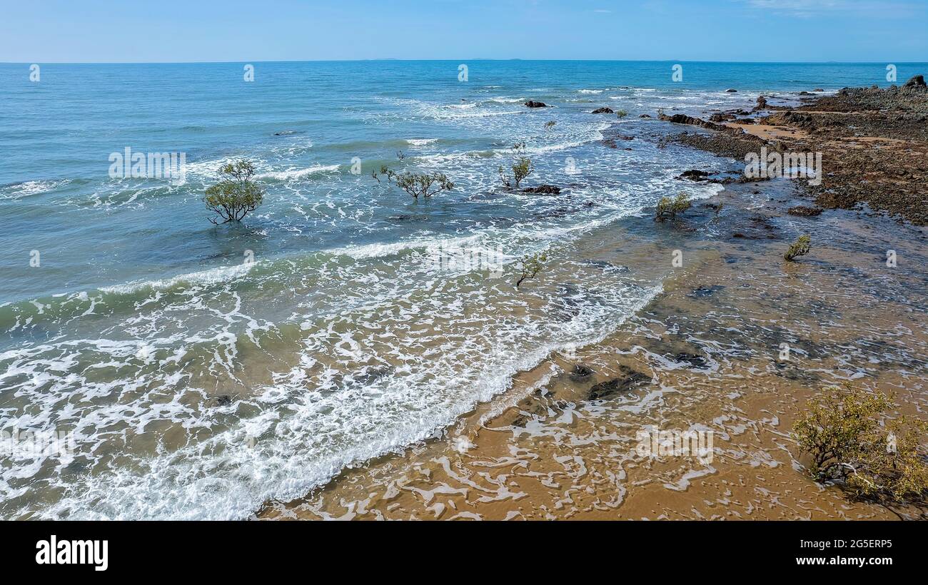 In volo su onde gelide verso rocce esposte su una spiaggia di sabbia sotto un cielo blu mentre la marea recede Capo Palmerston Australia Foto Stock