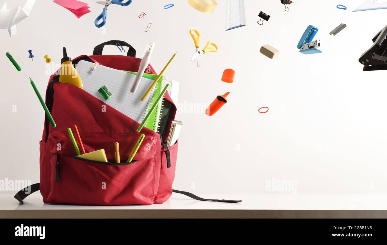 Scrivania di studio e forniture scolastiche che volano uscendo da uno zaino scuola con sfondo bianco isolato. Vista frontale. Composizione orizzontale. Foto Stock