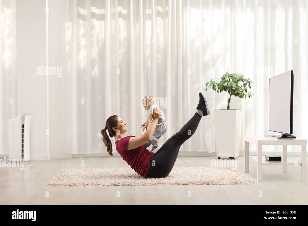 Giovane madre che si esercita sul pavimento e tiene un bambino a casa di fronte a una tv Foto Stock