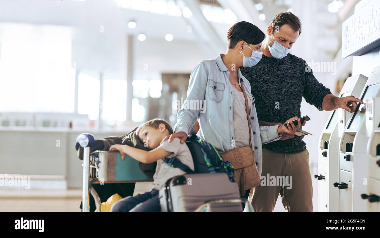 Accoppia usando un dispositivo di check-in self service con il loro bambino seduto sul carrello dei bagagli. Famiglia turistica che effettua il check-in all'aeroporto durante la pandemia. Foto Stock