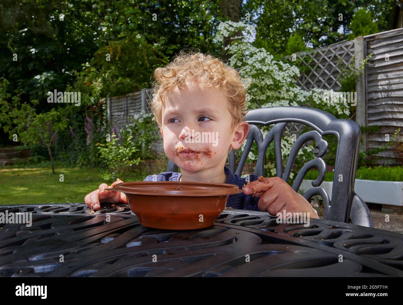 Un piccolo ragazzo caucasico bianco appiccicoso di quasi 3 anni con capelli biondi ricci si siede all'aperto con una bocca marrone gruby mangiare torta di cioccolato Foto Stock