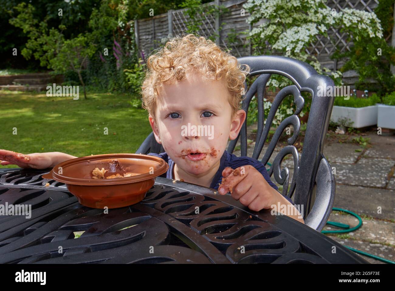 Un piccolo ragazzo caucasico bianco appiccicoso di quasi 3 anni con capelli biondi ricci si siede all'aperto con una bocca marrone gruby mangiare torta di cioccolato Foto Stock