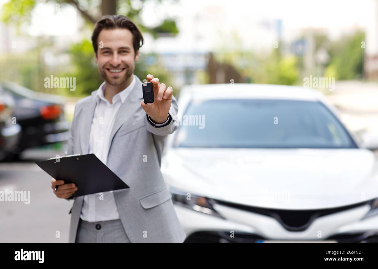 Agente di vendita offre al cliente di acquistare una nuova auto in distributore di assistenza. Sorridente giovane venditore di uomo bello in vestito dando la chiave al proprietario vicino nuovo au bianco Foto Stock