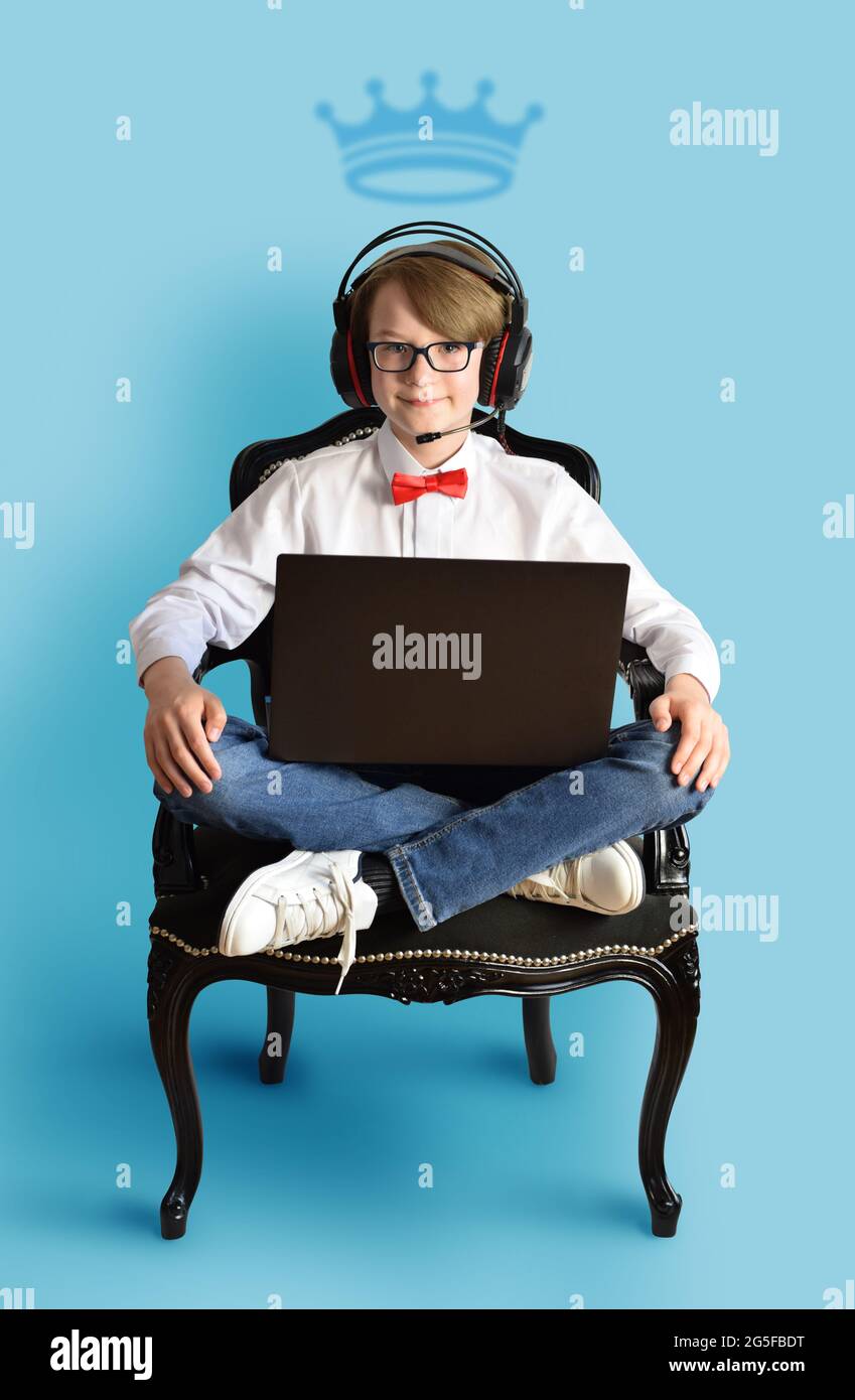 Ragazzo di 10 anni con laptop e cuffie seduti sulla sedia nera - lezioni online, concetto di e-learning, sfondo blu, Foto Stock