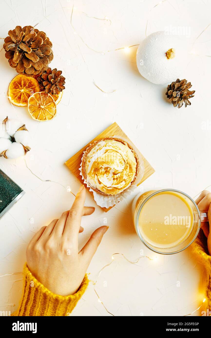 Posa piatta delle mani delle donne che toccano tazza di caffè e tartlet. Ragazza in maglione sta facendo colazione. Dolce dessert, bevanda, cono di pino e decorazioni su tavola bianca. Sfondo per le cartoline di festa. Foto Stock