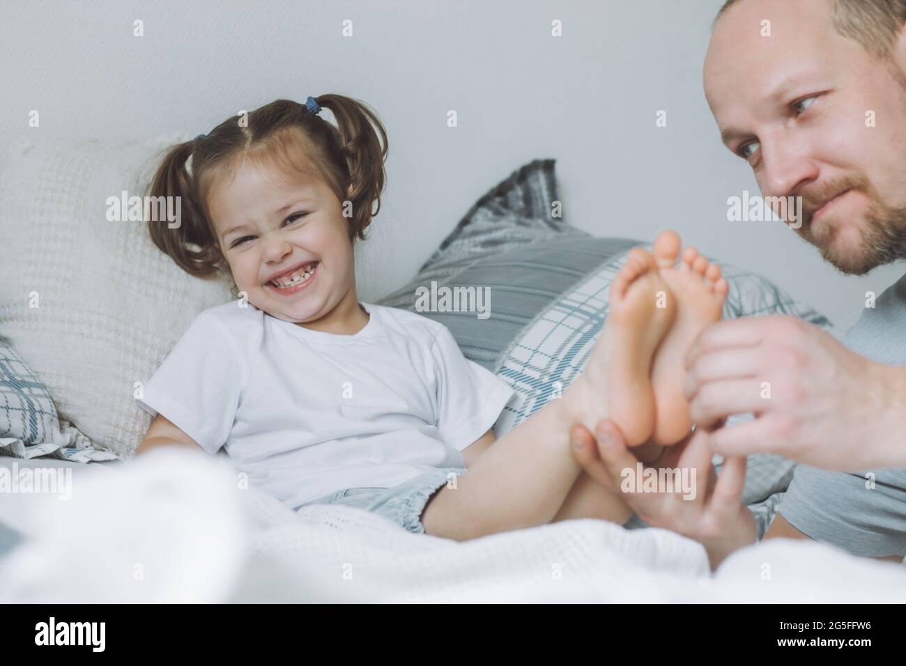 Padre gioca con la figlia piccola 2-4 a letto. Papà solletico i piedi dei bambini. Divertimento per tutta la famiglia Foto Stock