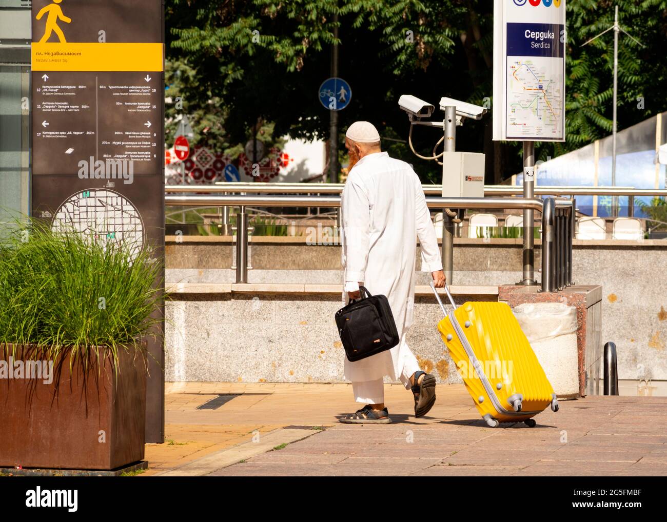 Uomo musulmano di mezza età che indossa un abito bianco e un cappuccio kufi con una cassa di lapton che tira la valigia gialla per le strade di Sofia, Bulgaria Foto Stock
