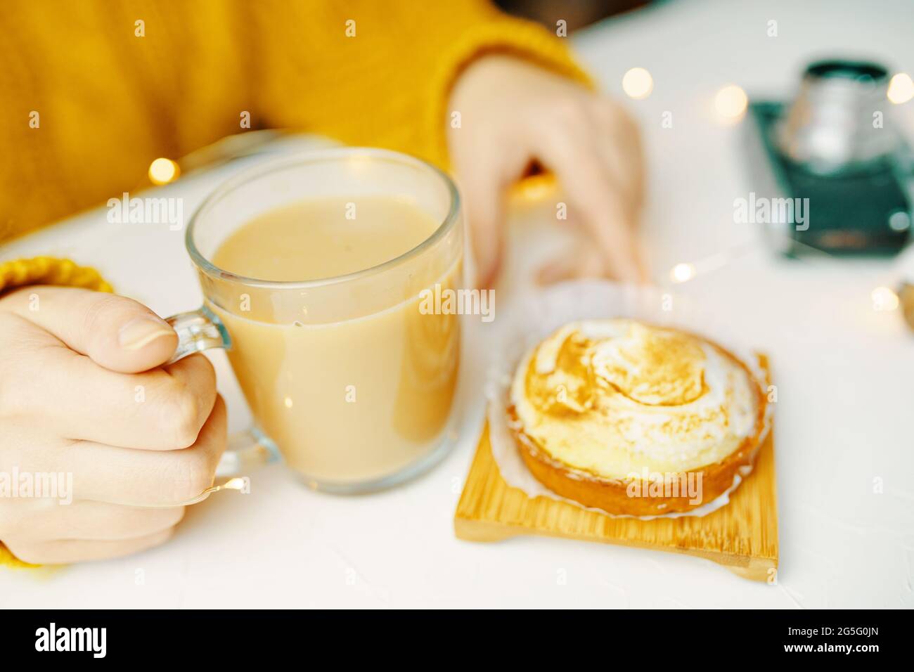 Le mani delle donne toccano tazza di caffè e tartlet. Ragazza in maglia maglione sta facendo colazione. Atmosfera romantica. Dolce dessert, bevanda rinfrescante e macchina fotografica sul tavolo bianco. Estetica della vita still. Foto Stock