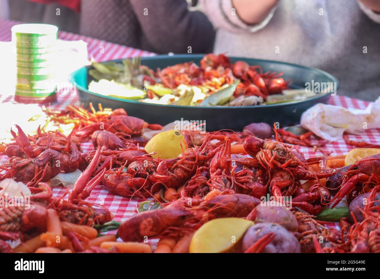 Crawfish bollito e verdure accatastate su tovaglia controllata rossa con vassoio di alimentazione e braccio di persona che mangia bokeh dietro - fuoco poco profondo Foto Stock