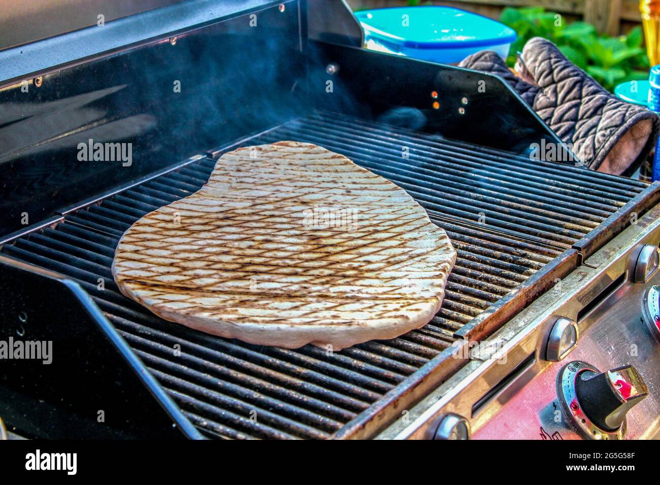 Rustica cucina casalinga di pane piatto su una griglia esterna con guanto da forno e plasticeria offuscata sul tavolo accanto ad esso - fuoco selettivo Foto Stock