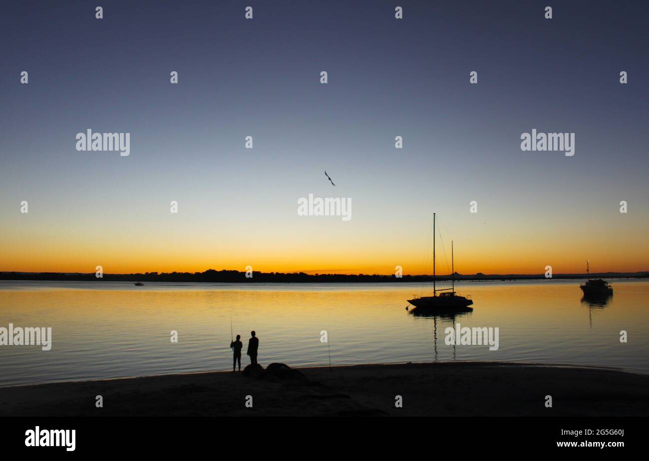 Silhouette di due pescatori a riva e barche a vela in acqua al tramonto Foto Stock