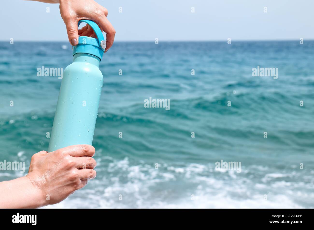 Bottiglia termica in acciaio nelle mani della donna in una giornata estiva soleggiata sul mare blu backgrond. Stile di vita sano, controllo dell'equilibrio dell'acqua, concetto di cura della pelle Foto Stock