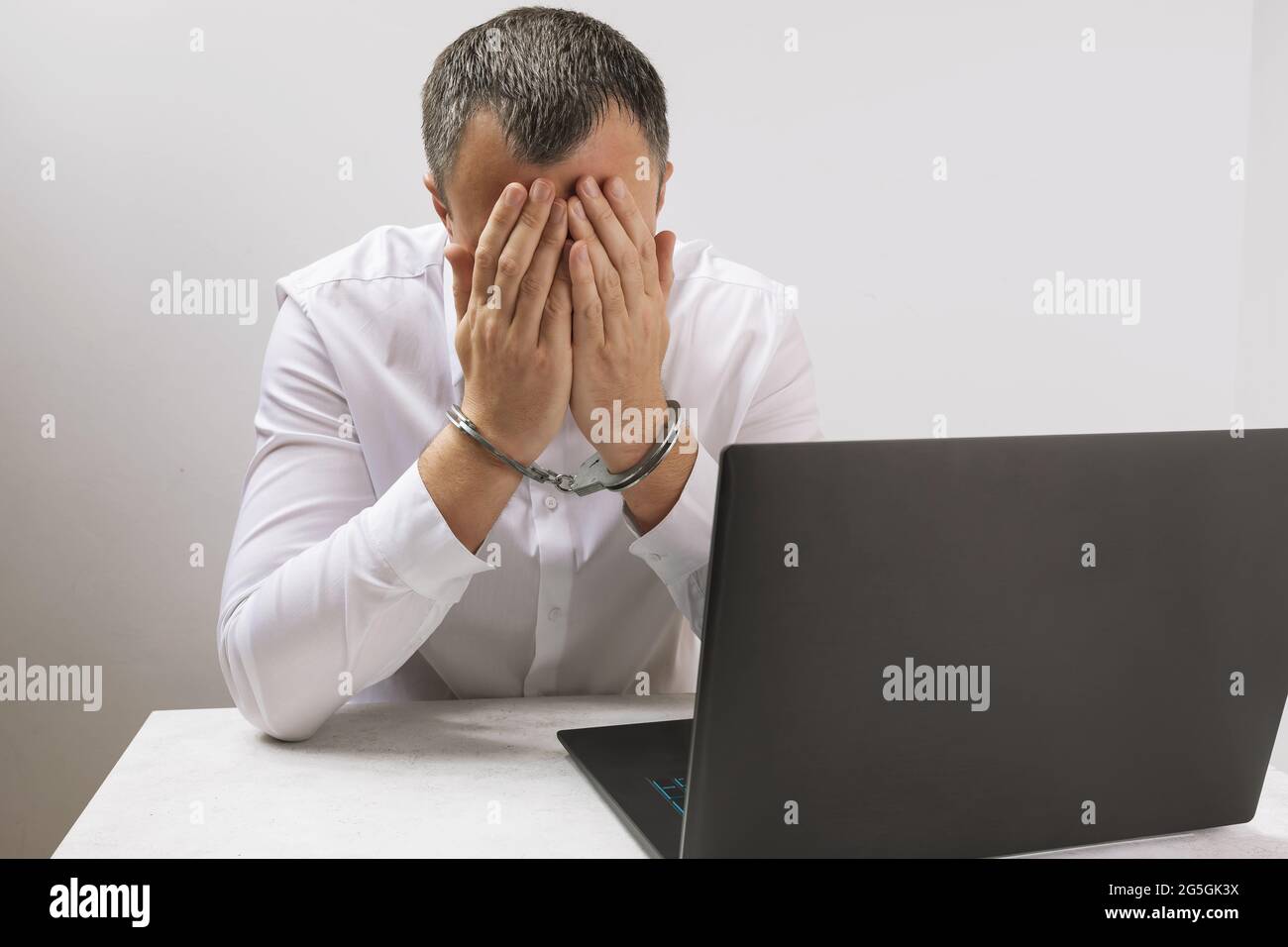 L'hacker catturato in manette vicino al portatile copre il suo volto con le mani, il concetto di lotta contro i crimini informatici Foto Stock