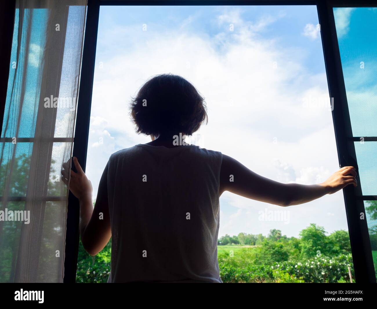 Immagine della parte posteriore della donna capelli corti che apre una grande finestra di vetro nella camera da letto al secondo piano della casa e che guarda al verde paesaggio e blu Foto Stock