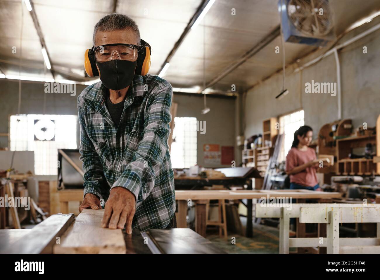 Falegname senior in maschera protettiva per il viso e cuffie che tagliano  lunghe assi in legno sul banco di lavoro Foto stock - Alamy