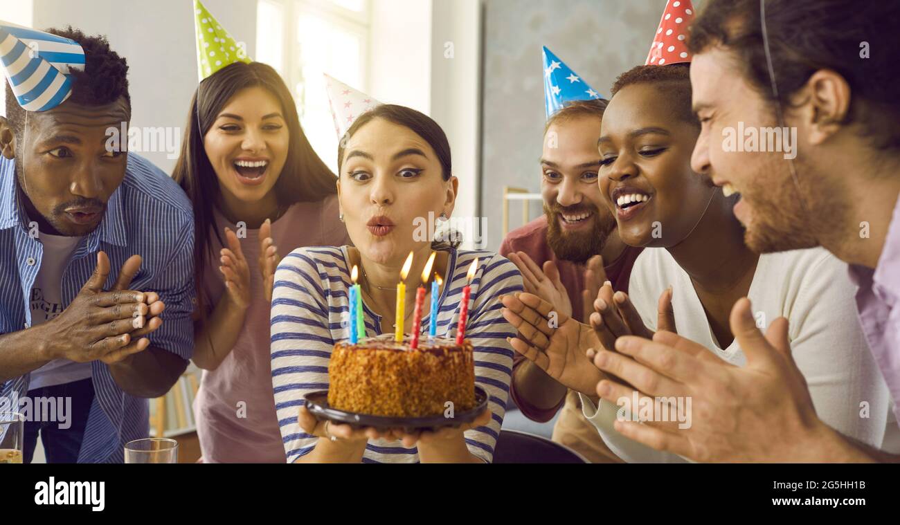 Gli amici applaudono come la ragazza di compleanno fa il viso divertente e soffia candele sulla sua torta Foto Stock