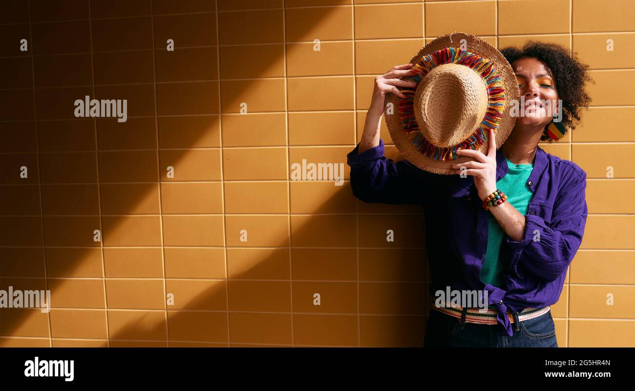 Giovane donna hipster multietnica con un cappello di colore paglierino nelle mani vicino a una parete urbana di piastrelle arancioni con ombra che cade dal sole Foto Stock