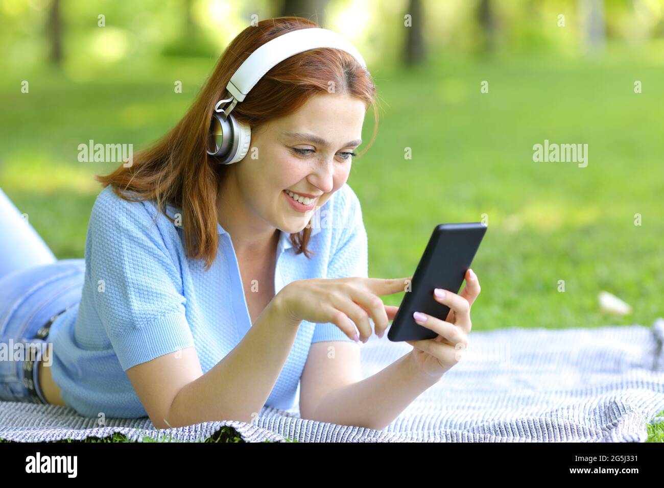 Donna felice che indossa le cuffie wireless che ascolta la musica controllando lo smartphone in un parco Foto Stock
