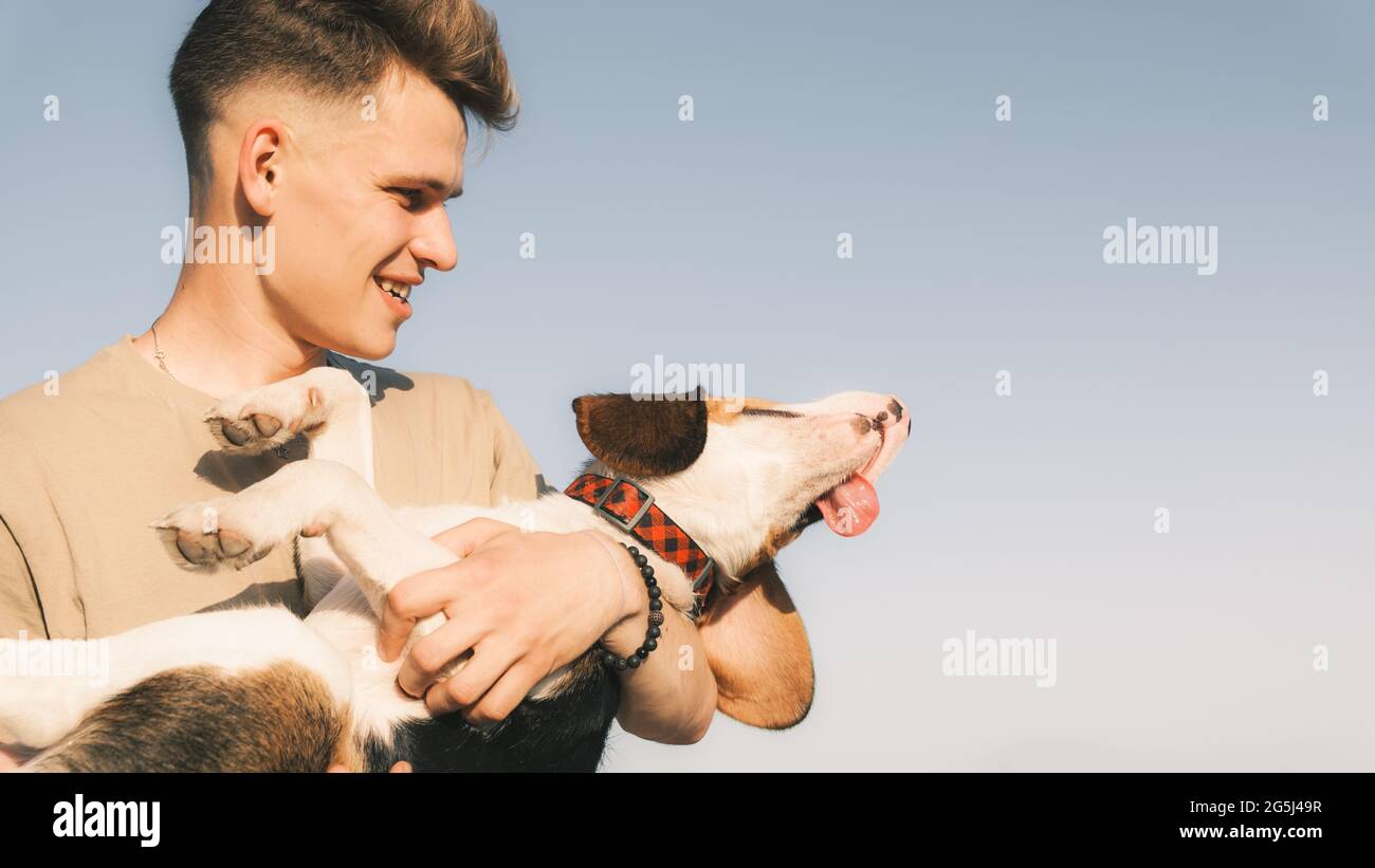 Il giovane uomo tiene il suo cane beagle felice in mani, sparato contro cielo blu chiaro con spazio di copia. Stile di vita con animali domestici, buon proprietario di cucciolo, cane purebred Foto Stock