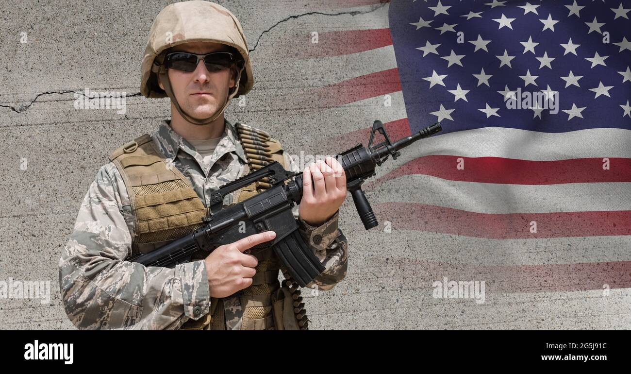 Composizione di un soldato maschio che indossa occhiali da sole che tengono una pistola, contro parete di cemento e bandiera americana Foto Stock