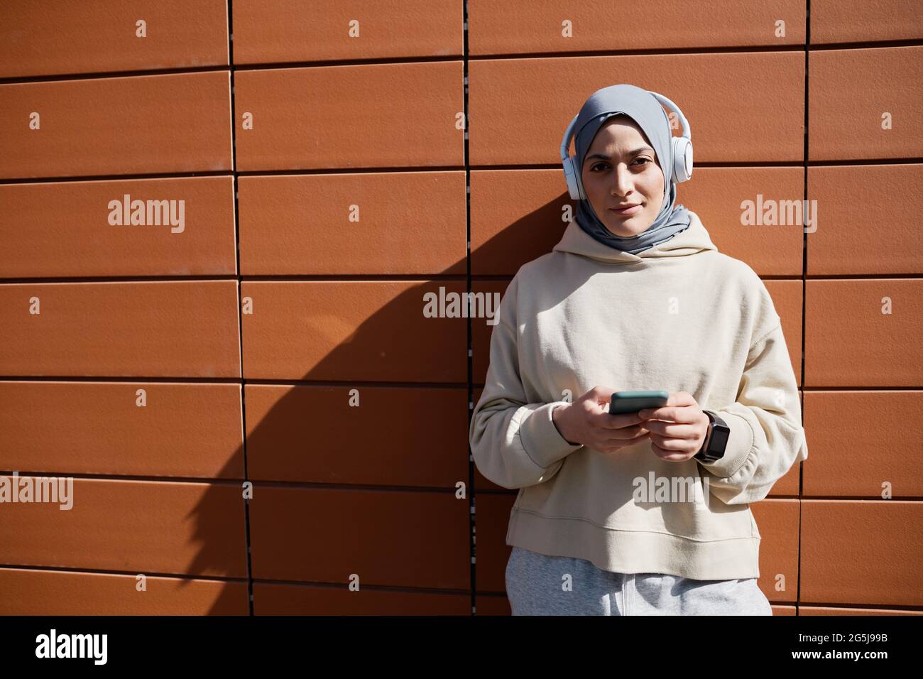 Ritratto grafico in vita di una donna moderna del Medio Oriente che utilizza uno smartphone in città illuminato dalla luce del sole e indossando cuffie, spazio per la copia Foto Stock