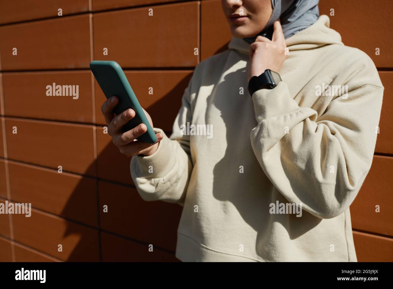 Ritratto troncato della donna medio-orientale contemporanea che tiene uno smartphone all'aperto illuminato dalla luce del sole mentre si trova accanto a una parete di mattoni, spazio per la copia Foto Stock