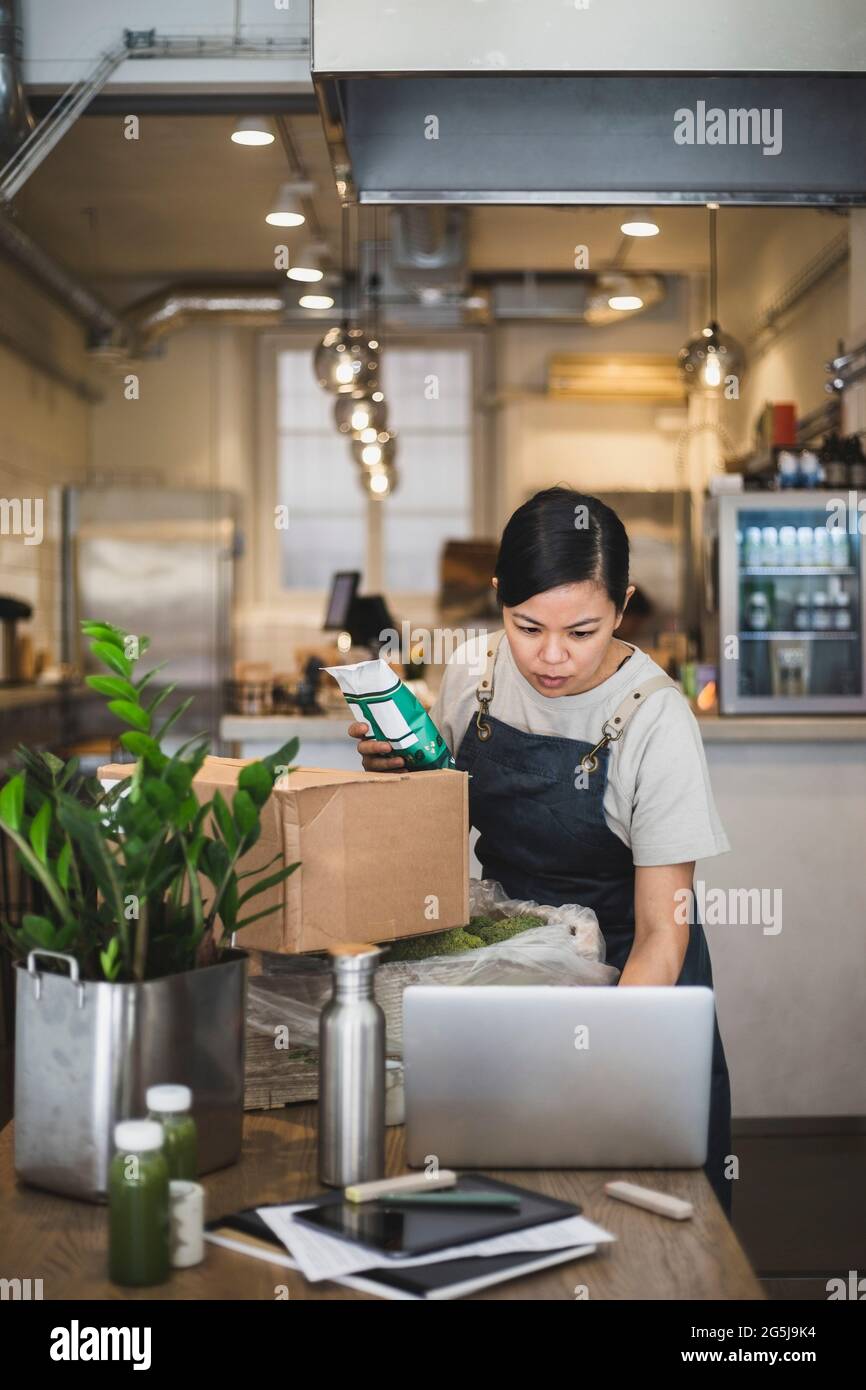 Dipendente femminile che lavora con un computer portatile mentre tiene il pacchetto di cibo in negozio Foto Stock