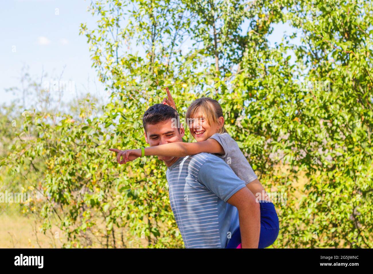 Fratello disinnescato che dà la sorella cavalcare sulla schiena. Ritratto di ragazza felice con corna divertenti sulle spalle dell'uomo, piggyback. Famiglia che gioca all'esterno. Estate Foto Stock