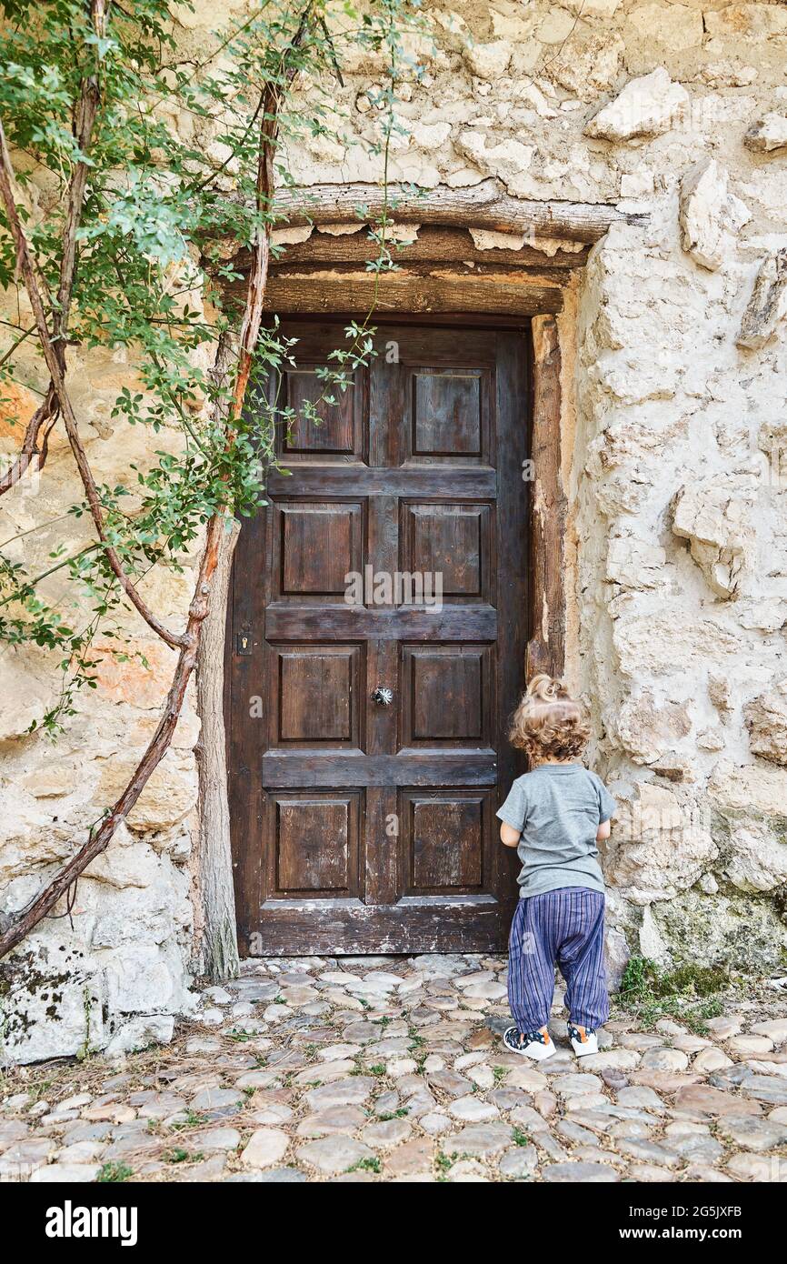 Vista posteriore di un solitario bambino che si trova all'esterno di fronte alla porta chiusa in legno di una vecchia casa. Concetto di solitudine. Fotografia verticale. Foto Stock